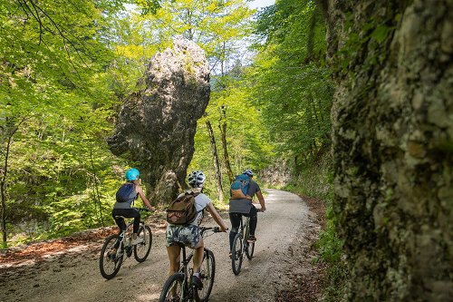 Kolesarjenje po Krajinskem parku Zgornja Idrijca, trije kolesarji na gozdni cesti pred pokončno kamnito strukturo Babji zob.